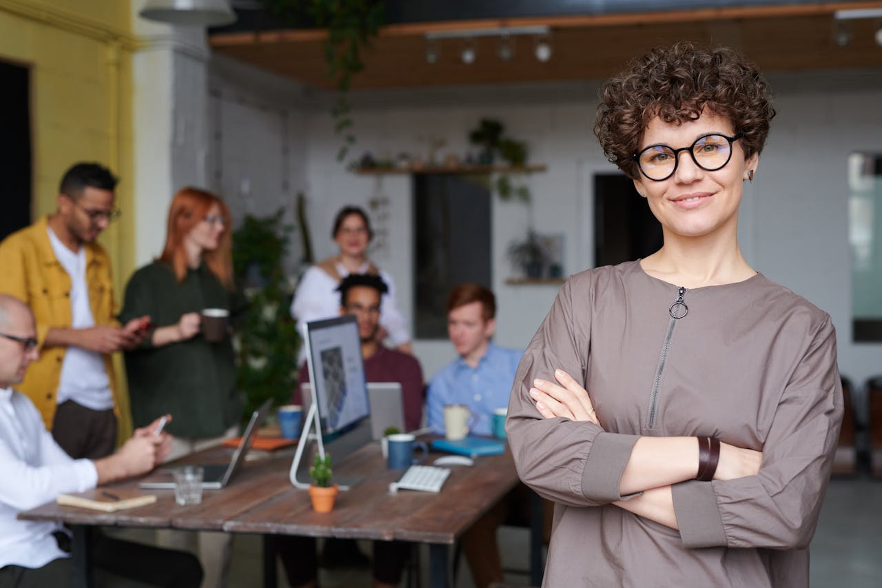 Services-02 Smiling businesswoman with curly hair stands confidently in a modern office space with colleagues.